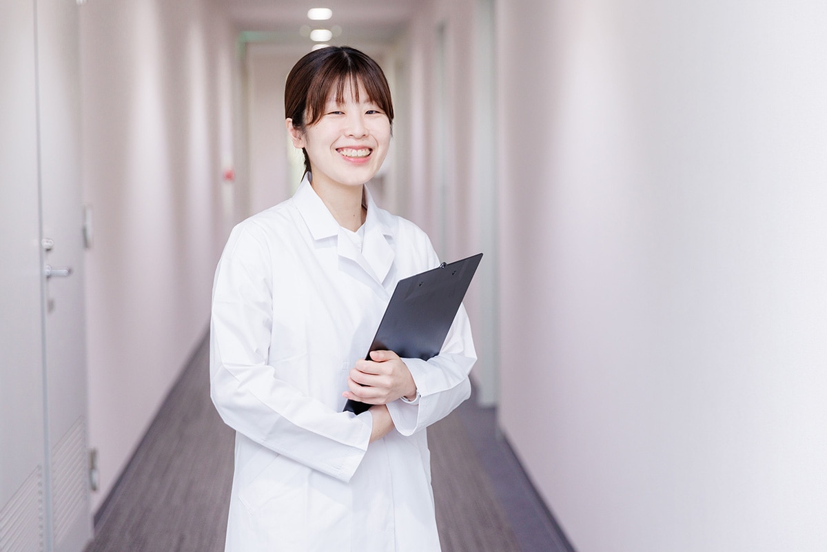 廊下で白衣を着てバインダーを持っている、治験コーディネーター（CRC）のOさん