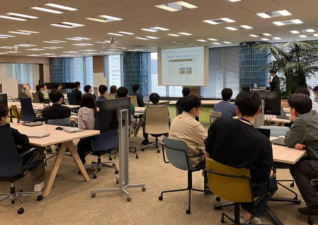 新入社員研修風景。セミナールームで多数の受講生がプロジェクターの画面を見ながら、集中して実習に取り組む様子。