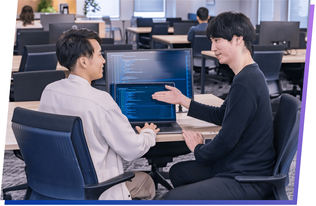 オフィスでの企業研修風景。講師が、プログラミングコードが映る画面を指し示しながら、生徒にマンツーマンで指導している様子。