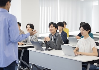 明るい研修室で、講師が数名の受講者に向けて立って説明している。受講者は各自のノートパソコンを使いながら熱心に耳を傾けている。