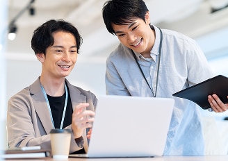 オフィスでノートパソコンを見ながら会話する二人の男性。AI開発の進捗を確認している様子。