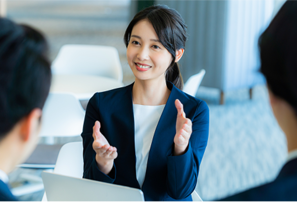 テーブルを囲んで座る複数の人物の中で、スーツを着た女性コンサルタントが身振り手振りを交えながら、明るい笑顔で相手に説明をしている。