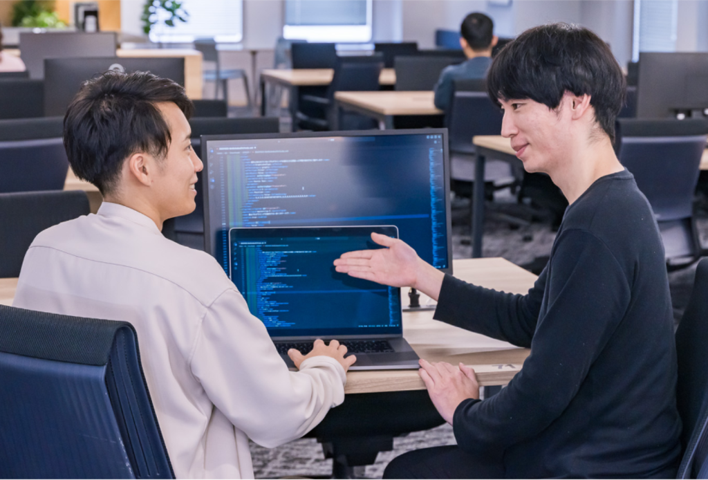 オフィスでノートパソコンと外部モニターに表示されたプログラミングコードを指差しながら話しているメンターと社員