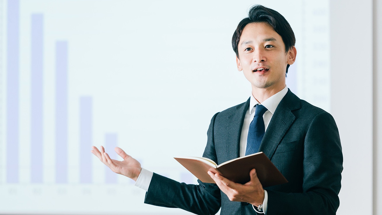 第一印象を高める自己演出研修イメージ。スーツ姿でノートを持ち、棒グラフを背景に信頼感を演出するプレゼンテーションの様子。