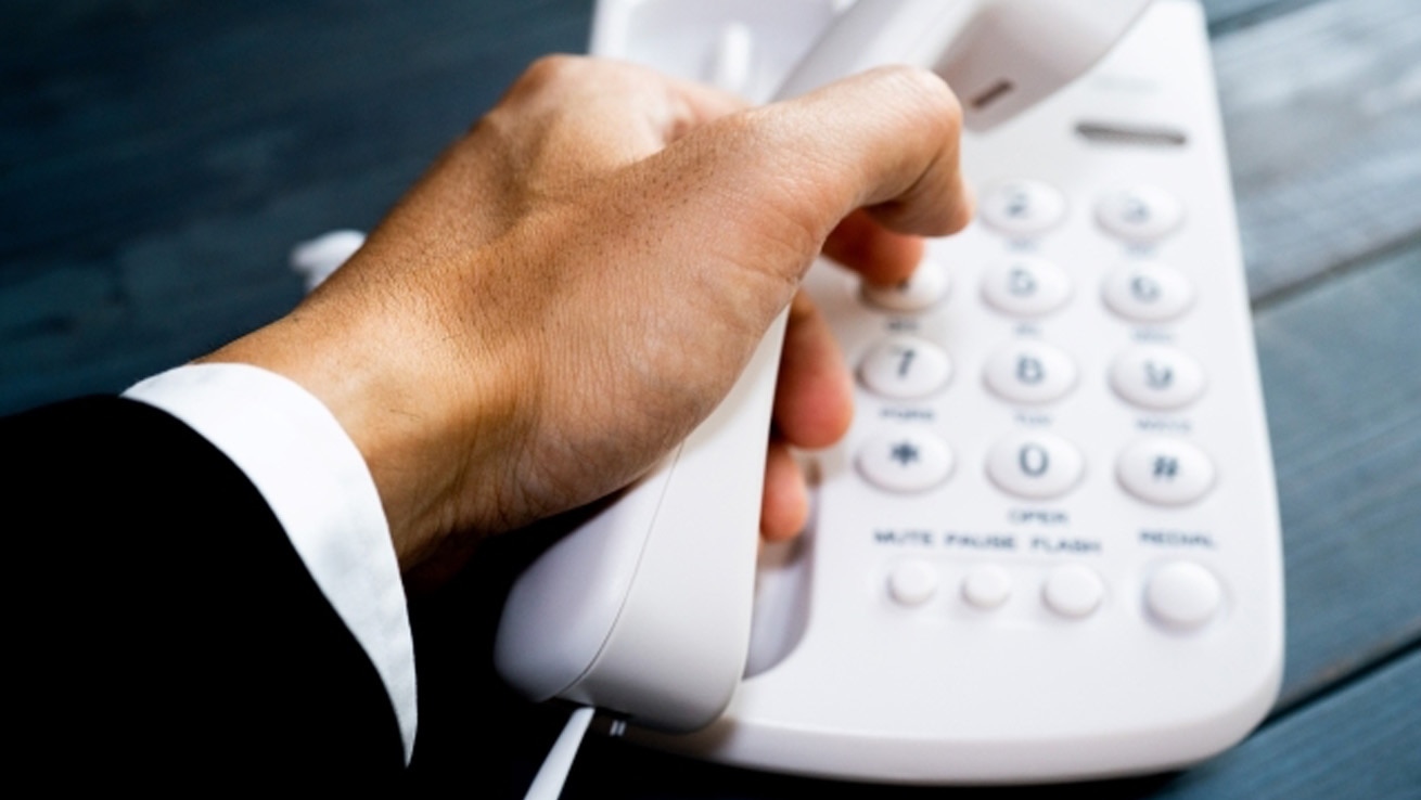 電話応対研修イメージ。ビジネスシーンで白い固定電話の受話器を取る手元、信頼感を伝える応対スキル習得を表現。