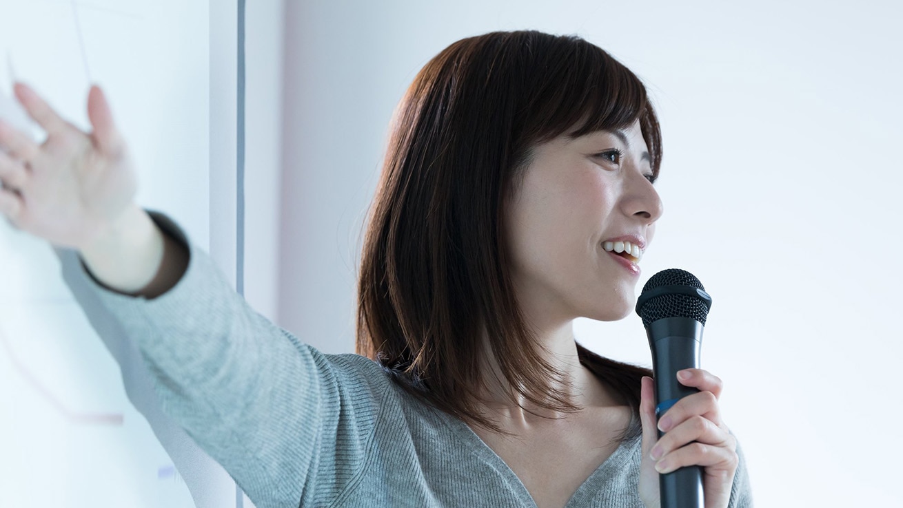 女性活躍推進研修 女性リーダー育成イメージ。マイクを持ち、前方に向かって説明する人物を通じて、リーダーシップや指導力を表現した写真。