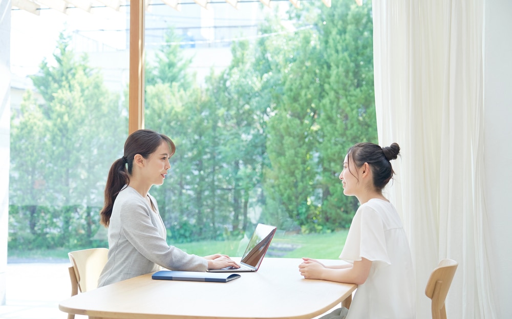 明るい窓辺のテーブルで、ノートパソコンを使う女性(左)が、向かいに座る女性(右)に何かを説明している。
