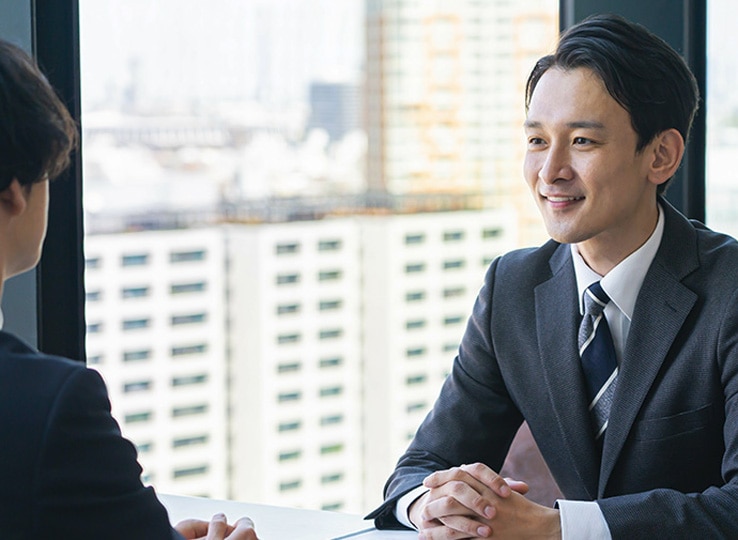 オフィスの窓際で、スーツ姿の2人がテーブルを挟んで向かい合い、ビジネスコミュニケーションを取っている様子。