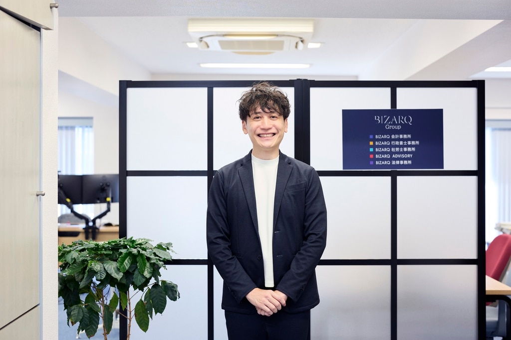 総合士業グループ「BIZARQ Group」のロゴパネル前に立つ吉岡和樹代表