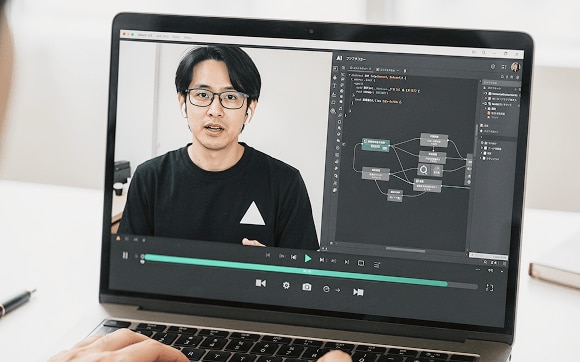 ノートパソコンの画面に映るオンライン動画講座の様子。画面左側には黒いTシャツを着て眼鏡をかけた男性講師が話し、右側にはAI構築用のフローチャートとコードが表示されている。