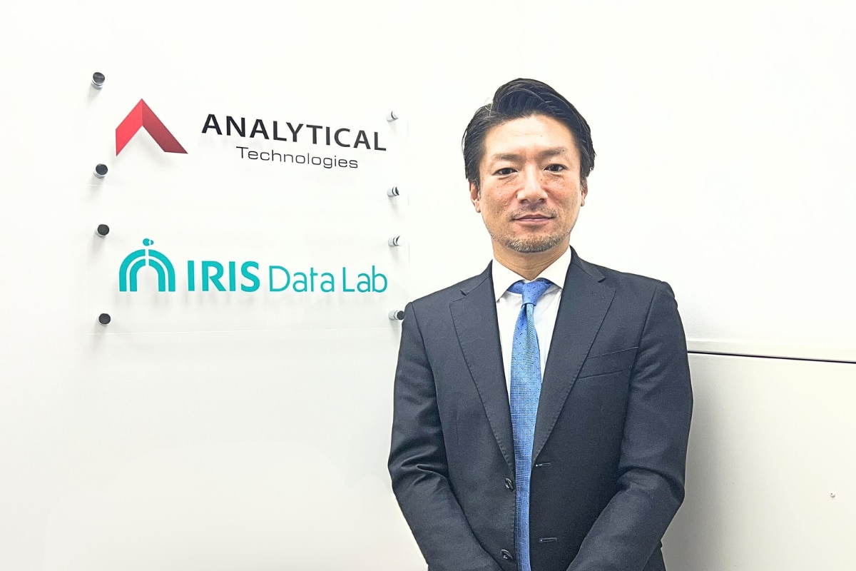IRISデータラボ株式会社のロゴ看板の横に立つ、スーツ姿の取締役・中塚様の全身カット