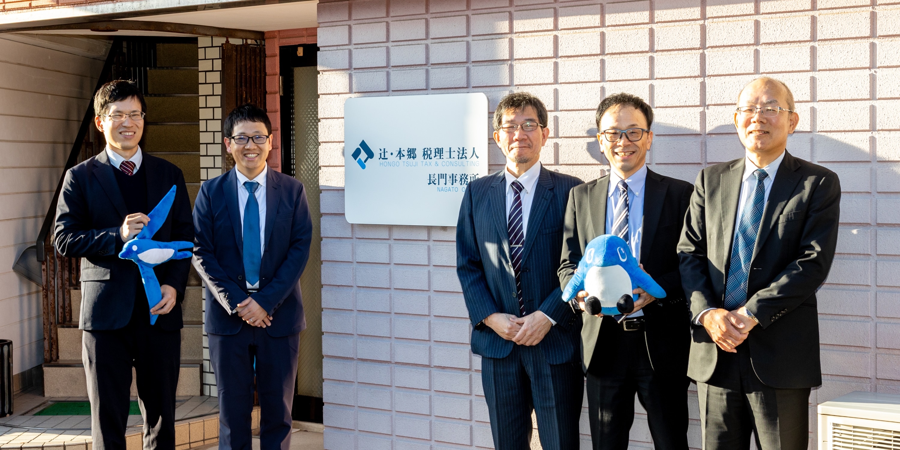 辻・本郷 税理士法人 長門拠点の看板前に立つプロジェクトメンバーの皆様。