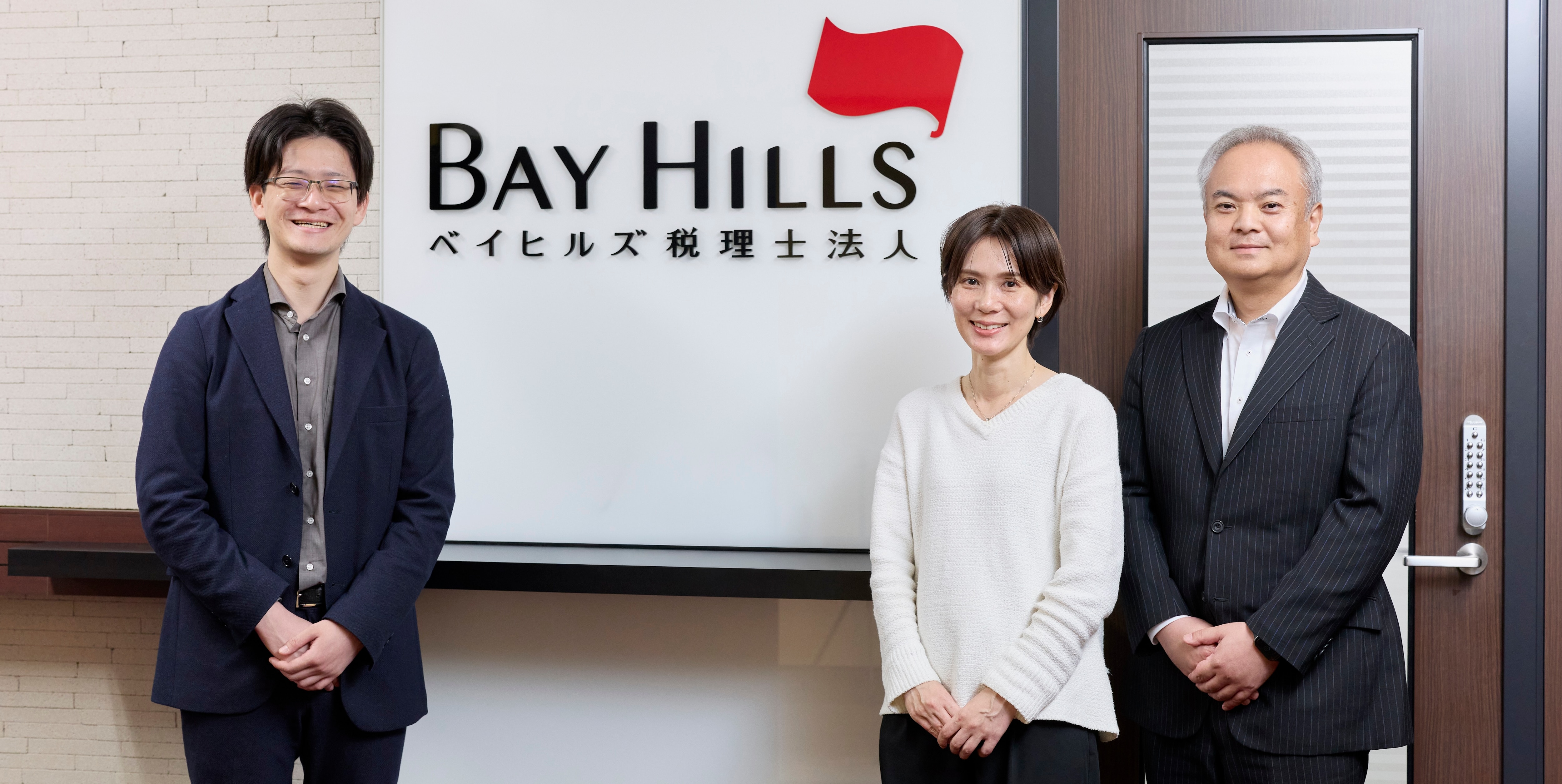 イヒルズ税理士法人のエントランスロゴ前に並ぶ木村氏、竹丸氏、加藤氏