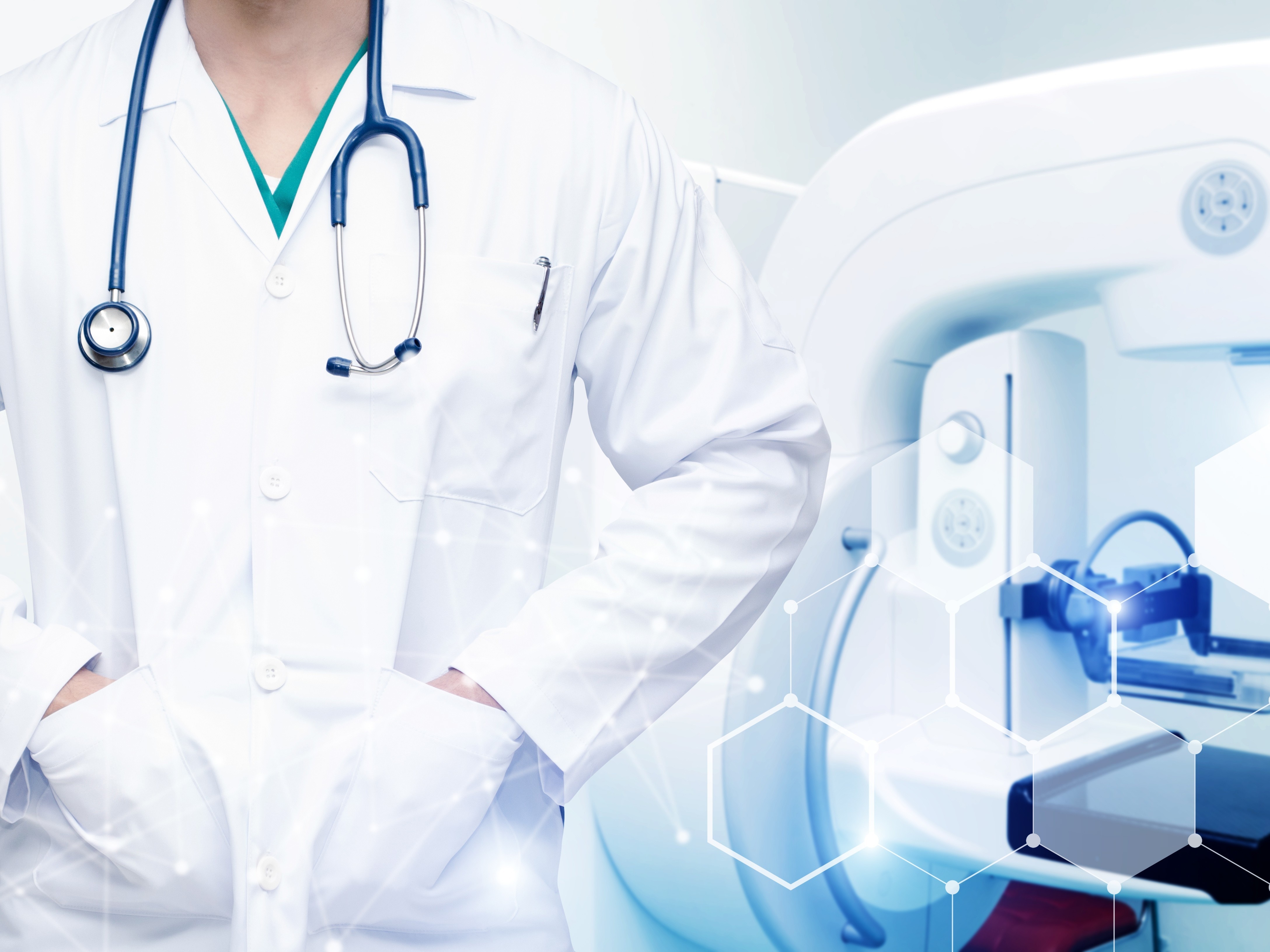 白衣を着た人物が首に聴診器をぶら下げている。後ろには大きな医療機器がある