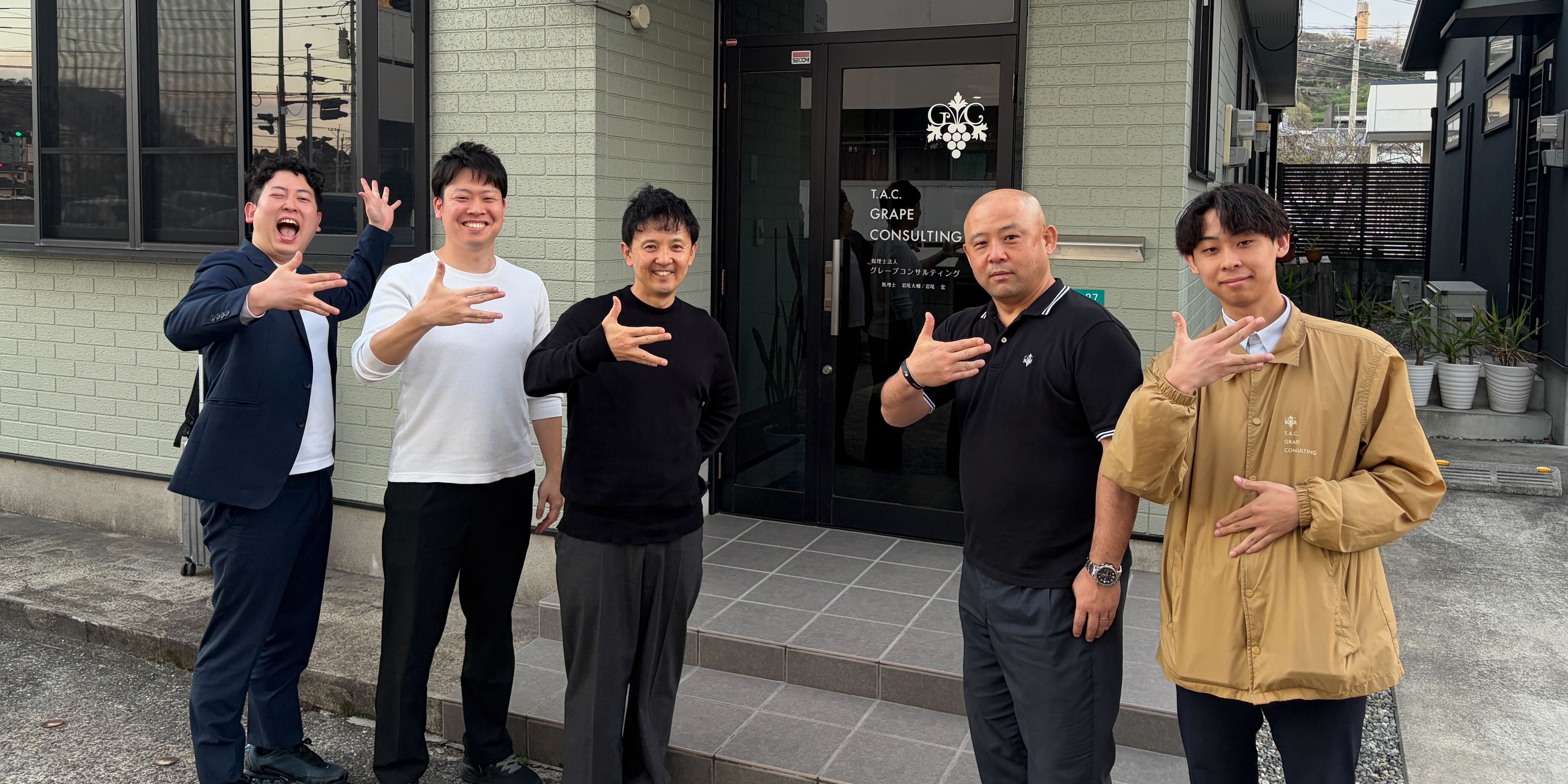 税理士法人グレープコンサルティングのオフィス前にて、ポーズをとる代表税理士の岩尾大輔氏（中央）、シニアマネージャーの岩尾英樹氏（右から2番目）、シニアスタッフの斉藤匠氏（左から2番目）ら